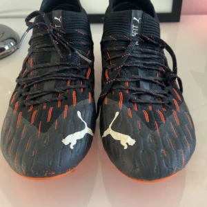 Svarta och orangea Puma fotbollsskor - Säljer ett par svarta Puma fotbollsskor med orangea detaljer och snörning. Skorna har en mönstrad ovansida och orange sula med dobbar. Puma-loggan syns tydligt på både fram- och baksidan. Perfekta för dig som vill sticka ut på planen.