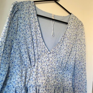 Ljusblå blommig långklänning med knappar - Söt ljusblå långklänning med små blommor i blå toner. Klänningen har v-ringning, långa ärmar och knappar framtill. Tillverkad i ett lätt och luftigt material, perfekt för vår och sommar.