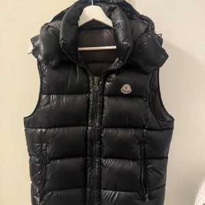 Svart dunväst från Moncler - Säljer en svart dunväst från Moncler med huva och dragkedja framtill. Västen har två fickor med dragkedja och Moncler-logga på bröstet. Perfekt för lager-på-lager och håller dig varm under kyliga dagar.