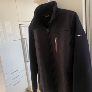 Svart jacka från Tommy Hilfiger - Säljer en svart jacka från Tommy Hilfiger med dragkedja framtill och hög krage. Jackan har diskret logga på bröstet och ärmen samt ett mönstrat tyg på axlar och bröst. Perfekt för kyliga dagar.
