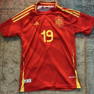 Spansk landslagströja Lamine Yamal 19 - Röd fotbollströja från Adidas med gula detaljer, nummer 19 och Lamine Yamal på ryggen. Officiell Spanientröja med landslagsemblem på bröstet och HEAT.RDY-teknologi. Klassisk design med gula ränder på axlarna.