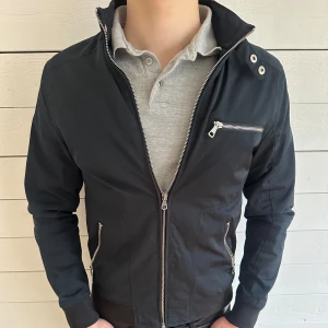 Svart jacka från River Island med dragkedjor - Säljer en svart jacka från River Island med flera silvriga dragkedjor och ståkrage. Jackan har lång ärm och en modern, stilren look som passar perfekt till vardags. Snygga detaljer med fickor och bröstficka med dragkedja.