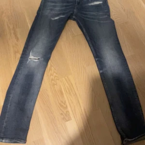 Mörkblå slitna jeans - Säljer ett par mörkblå jeans med slitningar på låret och knäet. Jeansen har en klassisk femficksdesign och raka ben. Perfekta för en avslappnad och trendig look.