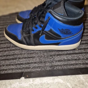 Nike Air Jordan 1 Retro High OG svart/blå - Säljer ett par Nike Air Jordan 1 Retro High OG i svart och blått med klassisk vinge-logga på sidan. Skorna har snörning, hög modell och vit sula. Perfekta för dig som gillar streetwear och sneakers med ikonisk design.