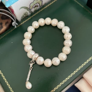 Thomas Sabo Pärlarmband - Ett vackert pärlarmband med en elegant berlock/charm från Thomas Sabo. Armbandet består av runda, vita skimrande odlade sötvattenpärlor. Pärlornas diameter är ca 1 cm. Berlocken är i 925 sterlingsilver och prydd med små, glittrande stenar samt en liten pärla längst ner. Perfekt för både vardag och fest. Fler charms kan fästas till. Storlek small, 15 cm, elastisk. Diameter ca 6-7 cm. Nyskick.