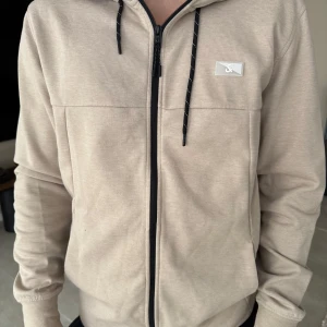 Beige hoodie med dragkedja - Säljer en beige hoodie från Sail Racing med svart dragkedja och svarta snören i huvan. Passar till allt och fantastik kvalité 