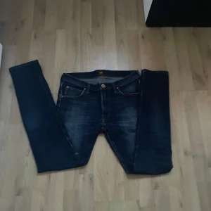 Mörkblå jeans från Lee Luke - Säljer ett par mörkblå jeans från Lee, modell Luke. Jeansen har klassisk femficksdesign, gylf med dragkedja och smal passform. Snygga slitningar framtill och Lee-logga bak på linningen. Perfekta för dig som gillar stilrena jeans!
