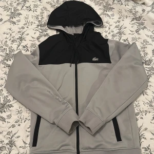 Grå och svart hoodiejacka från Lacoste - Säljer en snygg grå och svart hoodiejacka från Lacoste med dragkedja och huva. Jackan har logga på bröstet och Lacoste-tryck på ärmen. Perfekt för en sportig stil och har praktiska fickor med dragkedja.