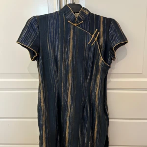 Mörkblå cheongsam-klänning med guldiga ränder från NONGHEMING - Snygg mörkblå cheongsam-klänning med guldiga vertikala ränder och dekorativa knappar framtill. Klänningen har korta ärmar, ståkrage och fina gulddetaljer längs kanterna. Perfekt för dig som vill sticka ut med en unik och elegant stil från märket YONGHEMINGYI i strl L