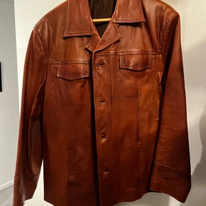 Brun skinnjacka med klassisk krage - Säljer en brun skinnjacka med klassisk krage och knäppning framtill. Jackan har två bröstfickor med lock och raka ärmar. Perfekt för dig som gillar tidlös stil och vill ha en jacka i äkta skinn. Vintage jacka med 20+ år med väldigt bra skick. Storleken sitter mellan M och L