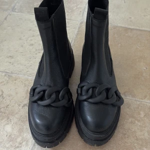 Svarta boots med kedjedetalj - Säljer ett par svarta boots med chunky sula och stor kedjedetalj framtill. Skorna har rund tå och är tillverkade i syntetmaterial med elastiska paneler på sidorna för enkel påtagning. Perfekta för dig som vill sticka ut med din stil.