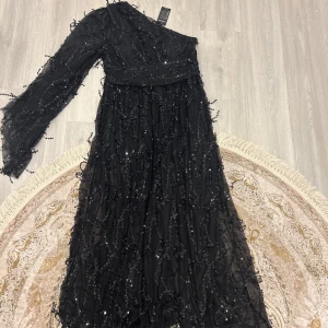 Svart långklänning med paljetter från Giffniseti - Säljer en svart långklänning från Giffniseti med glittriga paljettdetaljer och transparent mesh-tyg. Klänningen har lång ärm på ena sidan och är axelbandslös på andra, vilket ger en unik look. Perfekt för speciella tillfällen. Oanvänd. 