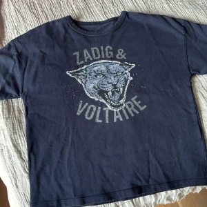 Mörkblå t-shirt från Zadig & Voltaire med tryck - Säljer en mörkblå t-shirt från Zadig & Voltaire med ett stort tryck av ett rytande kattdjur och text på bröstet. T-shirten har rund hals och korta ärmar. Perfekt för en cool och avslappnad stil.