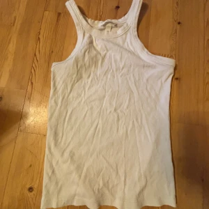 Vit ribbad linnetopp från H&M - Säljer en klassisk vit ribbad linnetopp från H&M. Toppen har ärmlös design och rundad halsringning, perfekt för varma dagar eller som lager under andra plagg.