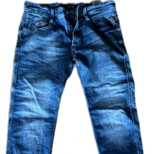Blå Replay jeansbyxor Anbass  31/32 - Säljer ett par klassiska blå jeans från Replay i modell Anbass. Jeansen har normal passform, fem fickor och dragkedja. Snygg tvätt med slitningar och raka ben. Perfekta för dig som gillar stilrena och bekväma jeans.