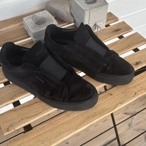 Svarta sneakers från Axel Arigato - Säljer ett par svarta sneakers från Axel Arigato i mocka med elastiskt band över vristen istället för snörning. Skorna har rund tå och platt sula, perfekt för en stilren och minimalistisk look.