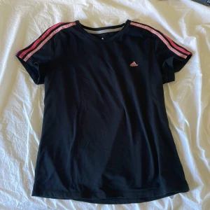 Svart t-shirt från Adidas med rosa ränder - Svart t-shirt från Adidas med tre rosa och röda ränder längs axlarna. Klassisk rund halsringning och korta ärmar. Liten Adidas-logga på bröstet. Perfekt för en sportig look.