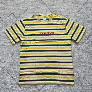 Randig t-shirt från Guess Jeans - Säljer en färgglad randig t-shirt från Guess Jeans med broderad logga på bröstet. T-shirten har gula, gröna, vita och lila ränder och är tillverkad i mjuk bomull. Perfekt för dig som gillar en retrostil med mycket färg.