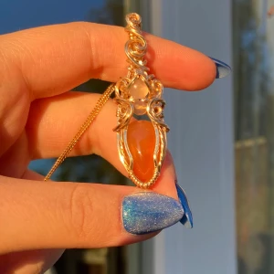 Guldberlock med orange droppformad sten - Vackert hänge i guldtonad metalltråd och två stenar, en orange och en ljusare ovanför. Hänget har snirkliga detaljer. Perfekt för dig som vill sticka ut med ett färgstarkt smycke.