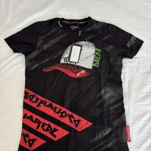 Svart t-shirt med tryck från Dsquared2 - Cool svart t-shirt från Dsquared2 med stort tryck av en keps och text i rött, grönt och vitt framtill. Unik design med grafiska detaljer och logga även på ärmen och ryggen. Perfekt för dig som gillar streetwear.