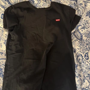 Svart t-shirt från Levi's - Svart t-shirt från Levi's med klassisk passform och liten röd logotyp på bröstet. T-shirten har rund halsringning och korta ärmar. Perfekt basplagg för garderoben.