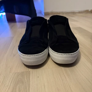 Svarta sneakers från Axel Arigato - Säljer ett par svarta sneakers från Axel Arigato med vit platt sula. Skorna är i mocka och har en stilren design utan snörning. Perfekta för dig som gillar minimalistisk stil och vill ha något som funkar till det mesta. Kontakta för mer information.