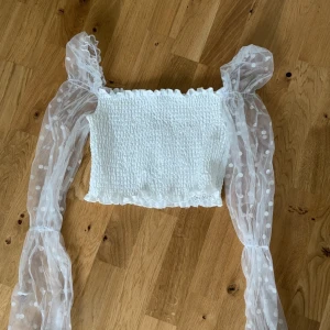 Vit croppad topp med puffärmar - Säljer en vit croppad topp med smockad byst och långa, transparenta puffärmar i mesh med prickigt mönster. Toppen har en fyrkantig urringning och är perfekt för en trendig look. Storlek s