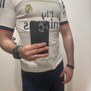 Real Madrid fotbollströja Ronaldo 7 - Säljer en vit Real Madrid matchtröja med svarta och guldiga detaljer. Tröjan har korta ärmar, Real Madrid-logga på bröstet, Fly Emirates-tryck och Ronaldo 7 på ryggen. Klassisk design med tunna ränder och broderade detaljer.