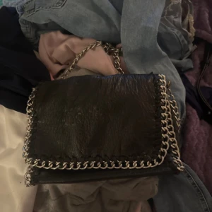 Svart axelväska med kedjedetaljer från Tiamo - Säljer en svart axelväska från Tiamo i skinnimitation med grova silverfärgade kedjor längs kanterna och som axelrem. Väskan har ett lock med magnetknapp och dragkedja inuti. Perfekt för dig som gillar edgy accessoarer.OBSSS!!! Vet inte om den är äkta köpte från secondhand och färgen börja ramla av