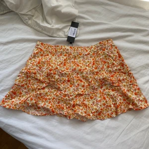 Blommig volangkjol från Motel - Supersöt kort kjol från Motel med blommigt mönster i orange, gult och vitt. Kjolen har volangdetaljer och dragkedja i sidan. Perfekt för sommarens alla tillfällen och lätt att matcha med en enkel topp.