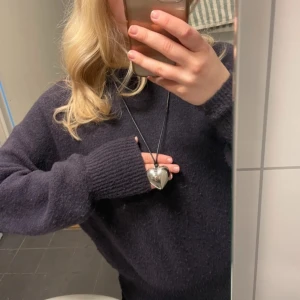 Halsband med silverfärgat hjärta - Säljer ett halsband med ett stort silverfärgat hjärta som hänge. Hjärtat hänger på ett svart snöre som kan justeras i längd. Perfekt accessoar för att ge din outfit en söt och stilren touch.