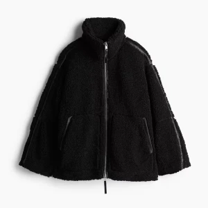 Svart teddyjacka med dragkedja - Mysig svart teddyjacka med hög krage och hel dragkedja framtill. Jackan har två stora fickor och en avslappnad passform, perfekt för kyliga dagar. Materialet är fluffigt och ger en trendig look.