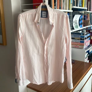 Ljusrosa linneskjorta från Zara - Säljer en stilren ljusrosa skjorta från Zara med klassisk krage och knäppning framtill. Skjortan är långärmad och tillverkad i ett mjukt bomullstyg. Perfekt för dig som gillar en clean och enkel look.