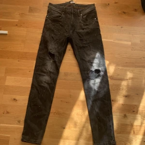Svarta slitna jeans med hål - Svarta jeans med raka ben och tydliga slitningar samt hål på båda benen. Jeansen har klassisk femficksdesign och små vita färgstänk över hela tyget. Perfekt för en avslappnad och edgy stil.