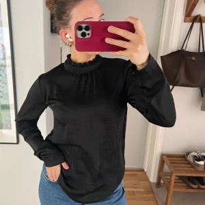 Svart blus med volangkrage från ICHI - Säljer en svart blus från ICHI med lång ärm och volangdetalj vid kragen. Blusen har en lätt glansig yta och en rak passform, perfekt att styla till jeans eller kjol. Klassisk och stilren modell med diskreta detaljer.