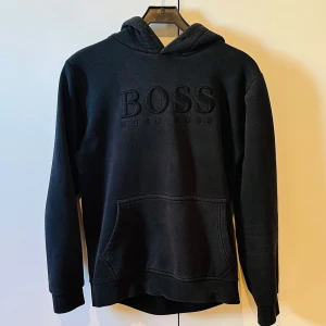 Svart hoodie från Hugo Boss - Säljer en svart hoodie från Hugo Boss med broderad logga framtill. Tröjan har huva, magficka och långa ärmar. Perfekt för en avslappnad och stilren look.