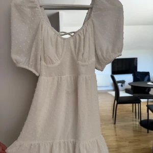 Vit kortklänning med puffärm och snörning i ryggen - Superfin vit kortklänning med puffärmar och söt struktur i tyget. Klänningen har v-ringning framtill och snygg snörning i ryggen. Perfekt för sommarens alla tillfällen!