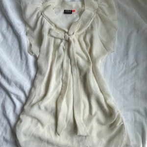 Vit blus med knyt från ONLY - Supersöt vit blus från ONLY med transparenta, luftiga korta ärmar och ett snyggt knyt framtill vid halsen. Blusen har en mjuk, flowy känsla och är perfekt för dig som gillar romantiska detaljer och en feminin look.