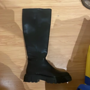 Svarta höga boots med grov sula - Säljer ett par svarta höga boots med chunky platt sula och rund tå. Skorna har dragkedja på insidan och är tillverkade i skinnimitation. Använda vid ett tillfälle, därav nyskick. Passar till både jeans och kjol.
