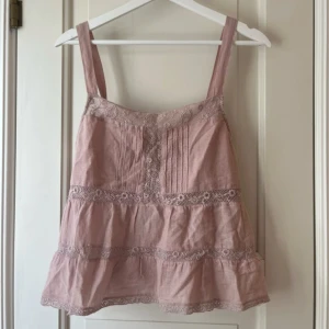 Rosa linnetopp med spets från Zara - Supersöt ljusrosa linnetopp från Zara med breda axelband och fina spetsdetaljer. Toppen har knyt i sidorna, dekorativa knappar framtill och volangkant nedtill. Materialet är linne och spets, perfekt för en chill sommardag. Passar dig som gillar romantisk och boho stil. Endast använt en gång