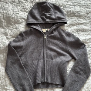 Grå croppad hoodie från Gina Tricot - Säljer en grå croppad hoodie från Gina Tricot med dragkedja framtill och huva. Tröjan har långa ärmar och en bred ribbad kant nedtill som ger en snygg passform. 