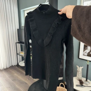 Svart polotröja med volang Second Female - Snygg svart polotröja från Second Female i storlek S. Tröjan har lång ärm, hög hals och en bred volangdetalj som går i V-form över bröstet. Materialet är stickat och känns mjukt mot huden. Perfekt för dig som gillar stilrena plagg med en twist.