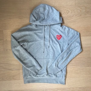 Grå hoodie Comme des Garçons Play - Grå hoodie från Comme des Garçons Play med ikoniskt rött hjärta med ögon på bröstet och stort hjärta på ryggen. Tröjan har huva med snörning, ribbade muddar och är tillverkad i mjuk bomull. Perfekt för dig som gillar streetwear med unik design.