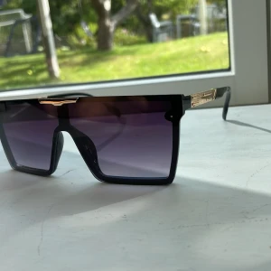 Svarta solglasögon med guld-detaljer - Säljer ett par riktigt snygga solglasögon med bred, rektangulär form och svarta bågar. Glasen är mörka med en lila ton och har coola guld-detaljer vid skalmarna och över näsan. Perfekt för dig som vill sticka ut med en modern och edgy look.