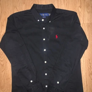 Svart skjorta från Ralph Lauren M - Klassisk svart skjorta från Ralph Lauren med vit knappstängning och röd broderad logga på bröstet. Skjortan har lång ärm, button-down krage och är i bomull. Perfekt för dig som gillar stilrena och tidlösa plagg med en touch av lyx.