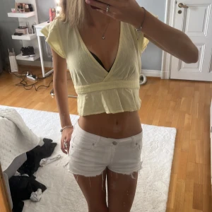 Ljusgul omlott blus med rosett - Säljer en ljusgul blus med omlott-design och v-ringning. Toppen är croppad med korta volangärmar och har en söt knytrosett bak. Materialet är lätt och luftigt, perfekt för varma dagar. Supersnygg till jeans eller shorts!