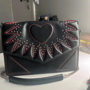 Svart axelväska med hjärtdetalj från Zara - Cool svart axelväska från Zara med ett stort hjärta och silvriga nitar på framsidan. Väskan har röda kontrastsömmar, geometriska former och en kedjerem i silver. Materialet är skinnimitation och insidan är fodrad med textil. Perfekt för att lyfta din outfit.