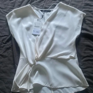 Vit omlottblus från Zara - Säljer en stilren vit omlottblus från Zara med knyt i midjan och v-ringad hals. Blusen har korta ärmar och är gjord i ett mjukt, glansigt material som ger en lyxig känsla. Perfekt för dig som gillar minimalistisk och chic stil.