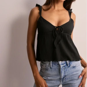 Svart linnetopp från Nelly - Svart linnetopp från Nelly med smala axelband och volangdetaljer. Toppen har en v-ringad hals och knyt framtill, samt lätt ribbad struktur. Croppad passform som ger en chill vibe och passar perfekt till jeans eller kjol. Använd fåtal gånger och i väldigt bra skick. Kan ta bättre bilder när de e ljust ute om de önskas💕