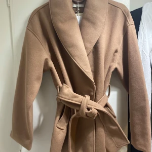 Beige kappa med bälte från Gina Tricot - Snygg beige kappa från Gina Tricot i storlek M. Jackan har bred krage, knytbälte i midjan och två fickor. Materialet är mjukt och känns lyxigt, perfekt för dig som gillar stilrena och klassiska plagg. Passformen är normal och ärmarna är långa. ALDRIG ANVÄND. 